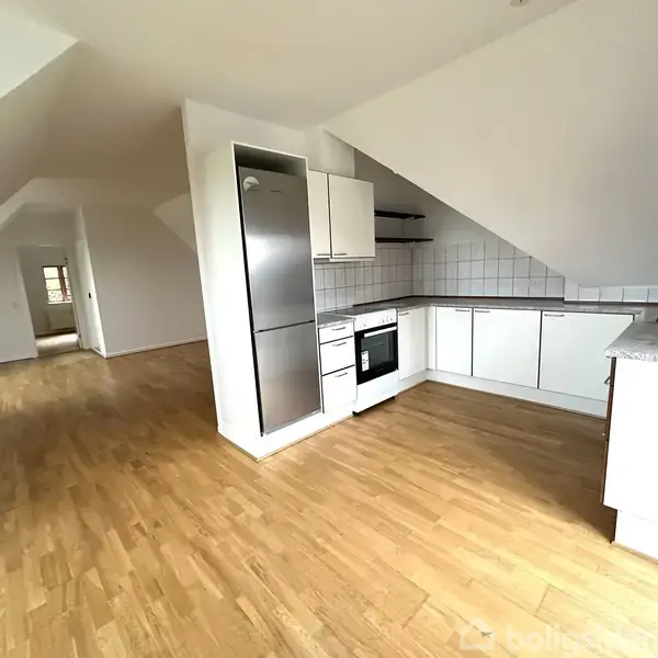 Et lyst køkken med hvide skabe og en stål-køleskab, placeret i en skråtagset lejlighed. Rummene har trægulve og en gang fører til andre værelser i lejligheden.