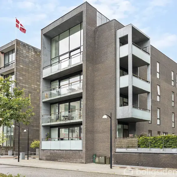 Moderne, firkantet lejlighedskompleks står stille, omgivet af et bymiljø. En rød-hvid dansk flag vajer fra taget. Fortov og gadehuse er synlige i forgrunden.