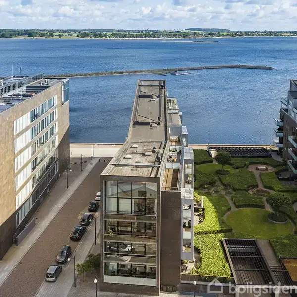 Flere moderne lejlighedsbygninger står langs en brostensbelagt vej ved en vandkant. Grønne haver og stier er synlige mellem bygningerne, med en blå himmel og havudsigt i baggrunden.