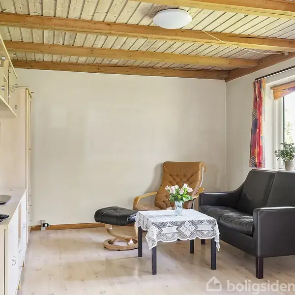Stue med sort sofa og lænestol med skammel placeret på trægulv. En buket blomster står på et lille bord mellem møblerne. Vindue med farverige gardiner til højre.