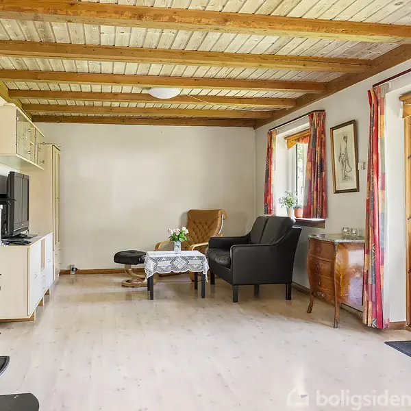 En stue med brændeovn i forgrunden, sofa og lænestol ved et lille bord, træloft og gardiner ved terrassen. Væggene har billeder, og et tv er placeret ved siden af skabene.