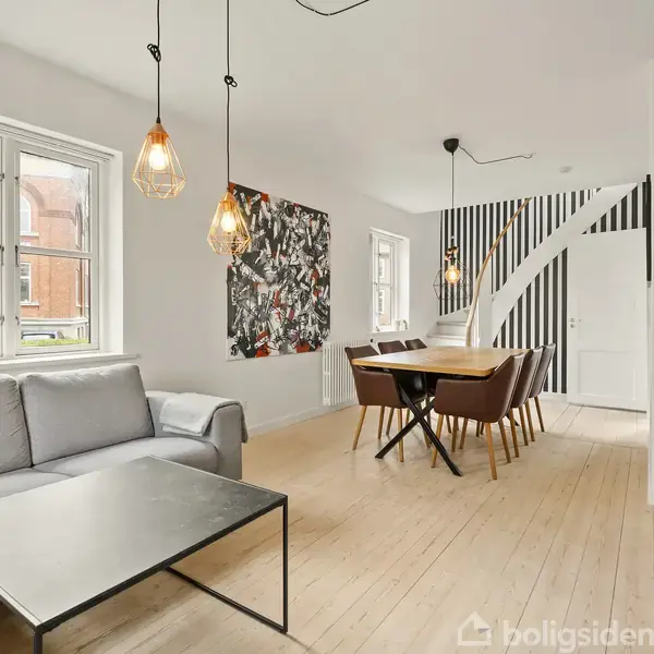 Grå sofa står ved et sofabord i en lyse, moderne stue med trægulve. Til højre ses et spisebord med stole, akkompagneret af dekorative lamper og en farverig vægdekoration.