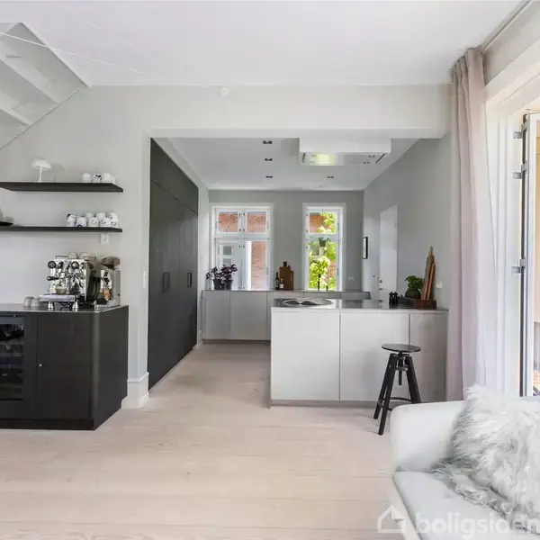 Køkken og stue i et moderne hjem. Sort skænk med kaffemaskine på venstre side, trappe ovenfor. Lys træbeklædning på gulvet, åbne hylder og lysegrå sofa til højre.