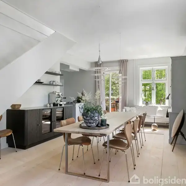 Spisebord med stole står i en moderne spisestue. Rummet har lyse gulve, store vinduer og en trappe, der giver adgang til en øvre etage. Dekorative elementer skaber en hyggelig atmosfære.