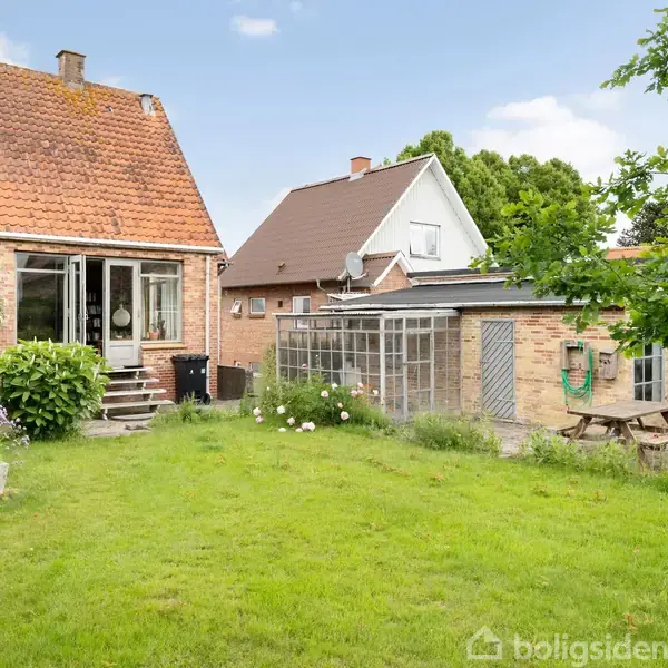 Et rødt murstenshus står stille i en grøn have med en lille terrasse og buske omkring. Der er et skur med et gyngestativ og et træbord ved siden af.