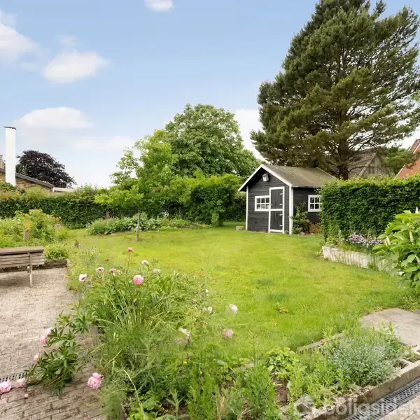 Et sort havehus står i en velplejet have med grøn græsplæne omgivet af buske og blomsterbede. I forgrunden er der en flisebelagt terrasse med en bænk og et drivhus.