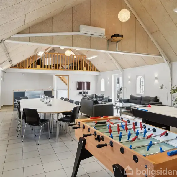 Et bordfodboldbord står uden aktivitet i et rummeligt opholdsrum med højt til loftet. Omkring er der et langt spisebord, sofaer, en plante og en trappe til en hems.