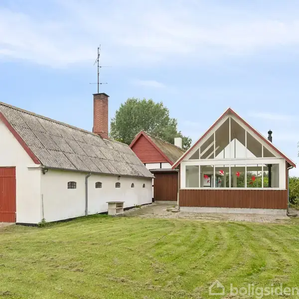 Et bindingsværkshus med stråtag og en moderne tilbygning med store glaspartier står ved en grøn græsplæne. Tilbygningen har en rødbrun facade, omgivet af træer og blå himmel.