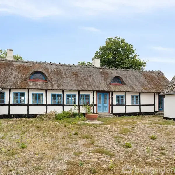 Bindingsværkshus med stråtag og blå vinduer står stille i en gårdsplads af ujævne brosten, omgivet af enkle, lavvandede bygninger og en stor grøn træ baggrund.