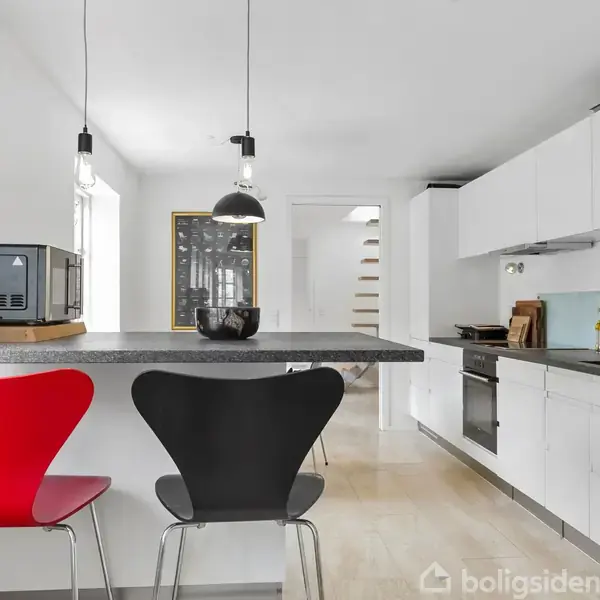 To røde og sorte stole står ved en køkkenbordplade. Ovenpå bordet står en mikrobølgeovn og skåle. Lyse, moderne køkkenskabe og apparater fylder rummet. Baggrund med hvide vægge og trappe.
