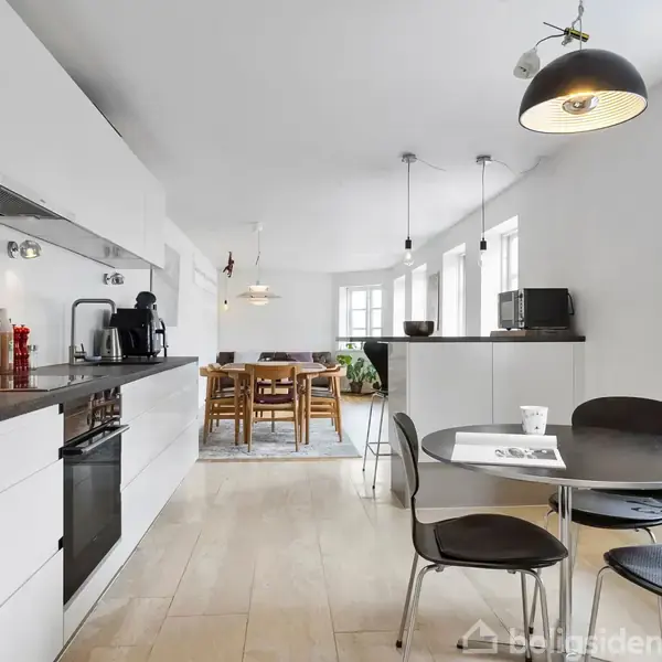 Køkken med hvidt interiør, sort bordplade. Rundt bord med aviser, stole omkring. Store vinduer, lamper hænger fra loftet. Baggrund viser spiseområde med træmøbler og tæppe.