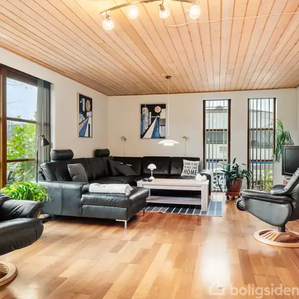 Sorte læderstole og sofa står i en stue med trægulve og træloft, omgivet af store vinduer, grønne planter og vægkunst. En lampe hænger over et sofabord i midten.