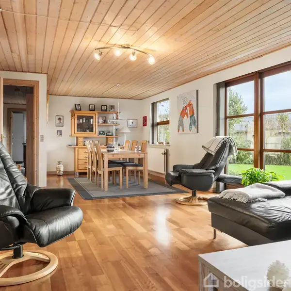 Sort læderstol står i et lyst stueområde med trægulv og træloft. Stuen har store vinduer, en spisebord i træ, grå sofa og indendørs planter, hvilket giver rummet en varm atmosfære.