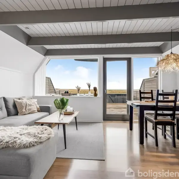 Et moderne stueområde med en grå sofa og et sofabord er placeret i lyse omgivelser. Spisebord med stole står ved siden af en glasdør, der fører ud til en altan med havudsigt.