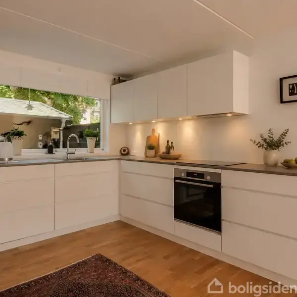 Et moderne køkken med hvide skabe og indbygget ovn. En bordlampe og planter tilføjer hygge, mens et stort vindue bringer naturligt lys ind. Gulvet er i træ.