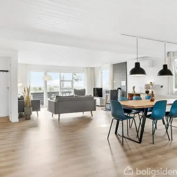 Et lyst og rummeligt stueområde med træslibegulv viser en spisebord med blå stole og en sofagruppe i baggrunden. Store vinduer bringer naturligt lys ind, og der hænger lamper over bordet.