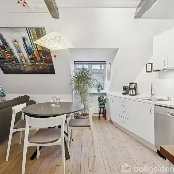 En moderne, lys stue-køkken-alrum med hvidt køkkenudstyr og trægulve. Et rundt spisebord med stole står foran en hvid væg, der har et stort farverigt maleri. En sofa ses til venstre.