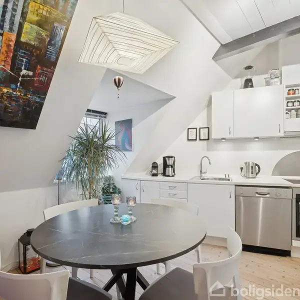 Rundt spisebord med stole placeret i et køkkenområde. Køkkenet har hvide skabe, opvaskemaskine, ovn og dekorationer som en stor plant, en maleri og en moderne loftslampe.