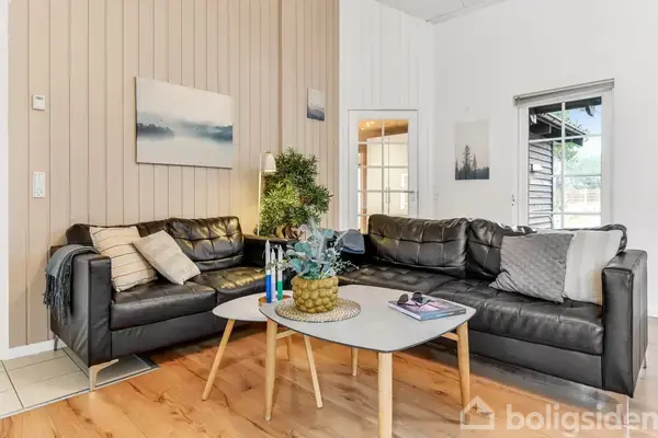 En sort lædersofa arrangeret i en vinkel står i en stue med trævægge. Et sofabord med pyntede detaljer står i midten. Bløde puder og kunst hænger på væggen.