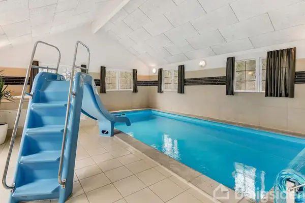 Blå rutsjebane står stille ved kanten af en indendørs swimmingpool. Rummet er lyst med hvide vægge og flere vinduer med mørke gardiner. Gulvet er flisebelagt.