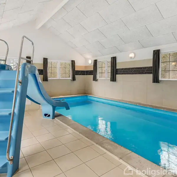 En indendørs swimmingpool med en blå rutsjebane ved siden, omgivet af hvide flisevægge og flere vinduer med gardiner. Poolområdet har en lys og ren atmosfære.