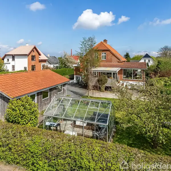 Et drivhus står i en have omkranset af grønne hække og træer. I baggrunden ses huse med røde og lyse tage under en blå himmel med skyer.