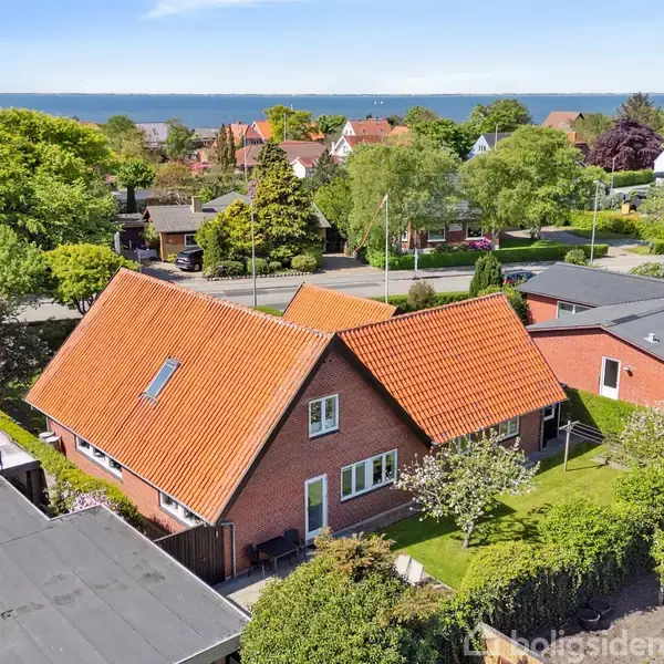 Et rødt murstenshus med rødt tag står i en velplejet have. Omgivet af andre huse ses en vej i et frodigt, grønt nabolag med udsigt til havet i horisonten.