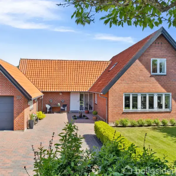 Et rødt murstenshus med en garage står stille på en brostensbelagt indkørsel. Det omkranses af en velholdt græsplæne og grønne buske under en blå himmel.