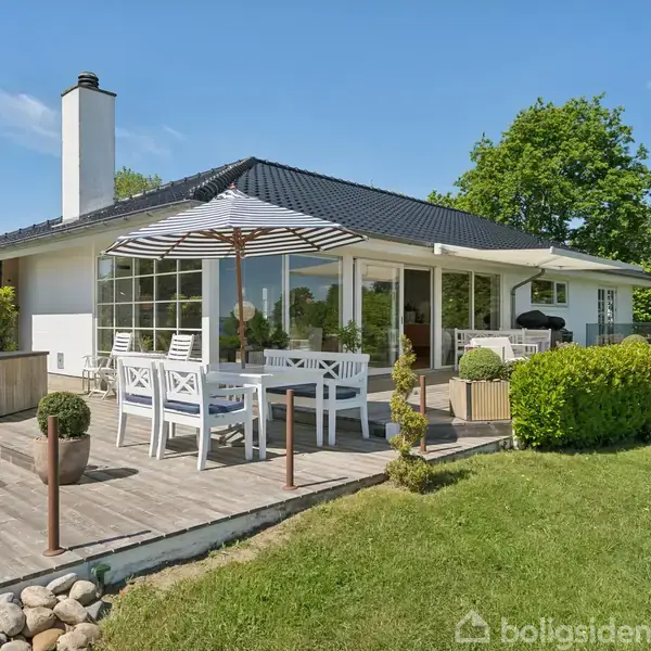 Et hvidt hus med store vinduer og sort tag står i en frodig have. Terrassebord med stole og stor parasol på trædæk. Grønt græs og buske omgiver området.