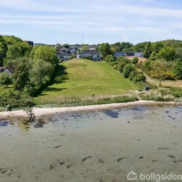 En stor græsplæne skråner ned fra en bolig omgivet af træer mod en stenstrand og klart vand. Huset ligger i en fredelig, grøn landskabsomgivelse.