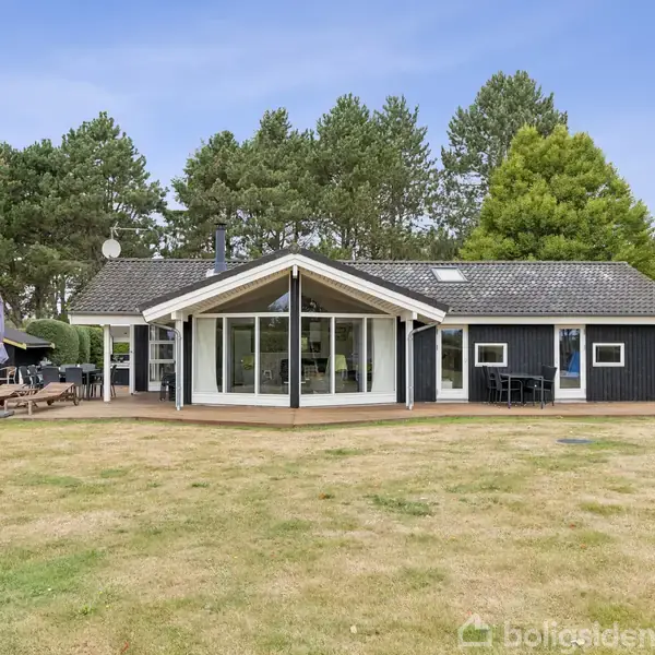 Et sort træhus med store vinduer står stille i en skovomgivet have på en solrig dag. Det har en terrasse og solstole ved siden af.