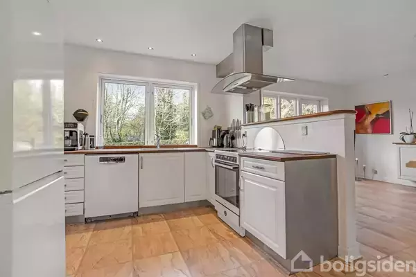 Et moderne køkken med hvide skabe har en ovn og komfur i midten. På bordpladen står køkkenredskaber. Store vinduer lader naturligt lys ind. En farverig maleri hænger på væggen.