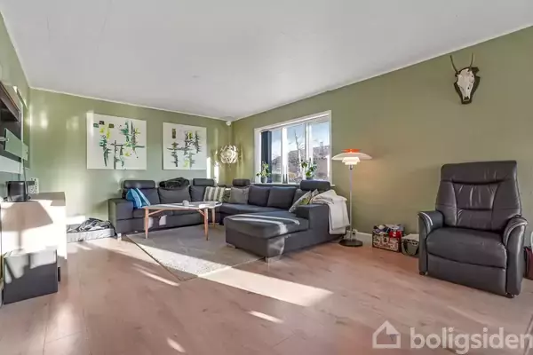 Stue med en mørkegrå hjørnesofa står ved et vindue, oplyst af dagslys. På væggene hænger kunst og et hjortegevir. Rummet har lyse trægulve og en gulvlampe.