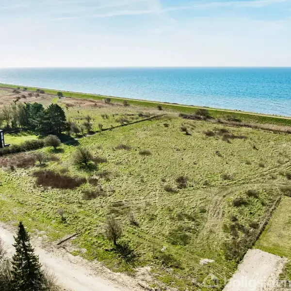 Grøn mark strækker sig mod havet, omgivet af få træer og buske. En enkelt bygning ses nær venstre side, mens kystlinjen strækker sig langs horisonten.