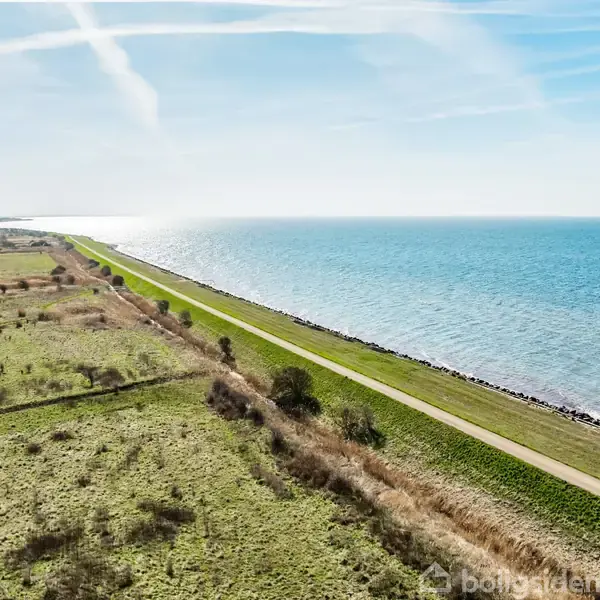 En smal vej følger en kystlinje ved siden af en græsmark, hvor havet strækker sig til højre under en klar, blå himmel.