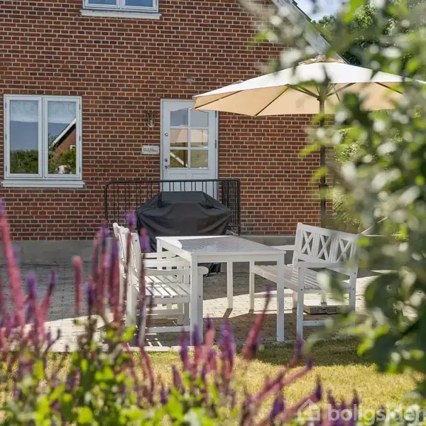 Et hvidt havesæt med parasol står på en terrasse foran et rødt murstenshus. Omkring terrassen ses grønne planter og lilla blomster. Ingen tekst er synlig.