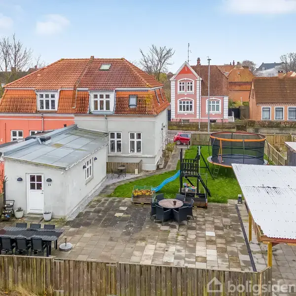 Et rødt tegltagshus med en legeplads og terrasse i baghaven. På terrassen står et bord og stole, omgivet af et træhegn i et boligområde.