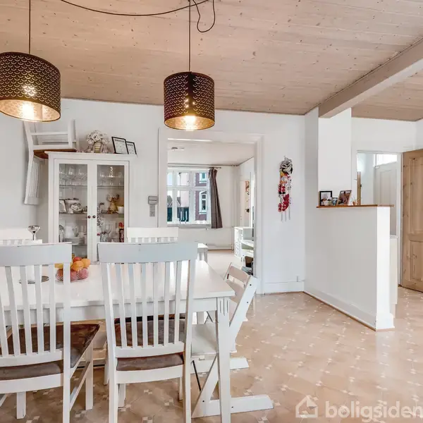 En spisestue med et hvidt spisebord og fire træstole. To sorte lamper hænger over bordet. På væggen til venstre ses familiefotos og teksten: "In this house, We are family".