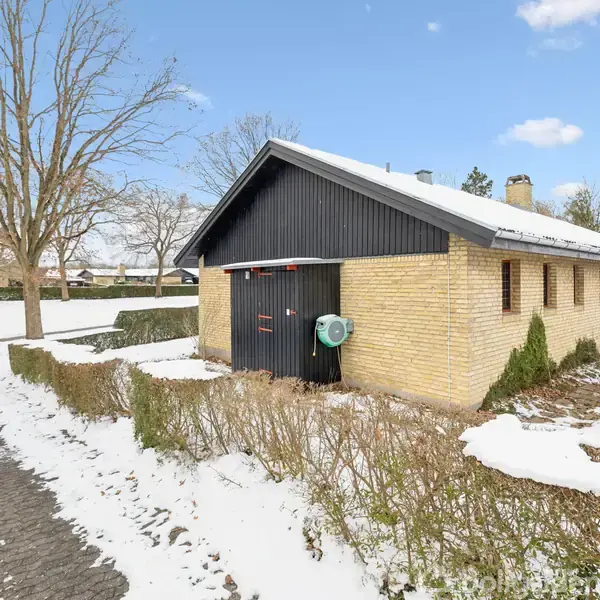 Et gulstenshus står stille i et snedækket landskab omkranset af bar træer og buske. Sneen dækker jorden og skaber en vinterlig atmosfære omkring bygningen.