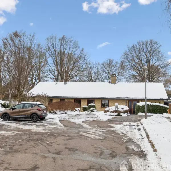 En brun bil holder parkeret foran et hvidt murstenshus med sne på taget. Omgivet af sneklædte træer og en overskyet himmel. Parkering på en halvrevet asfalteret vej.