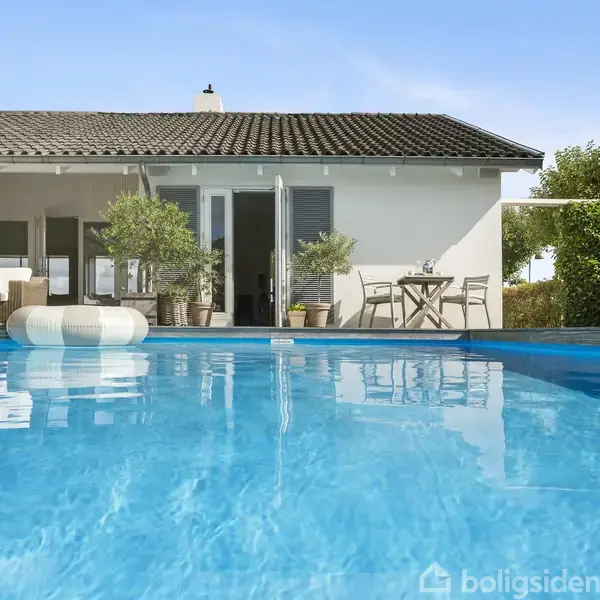 Swimmingpool med klart blåt vand foran et enkelt hvidt hus. I baggrunden ses havemøbler, planter i krukker og franske døre, der åbner mod terrassen.