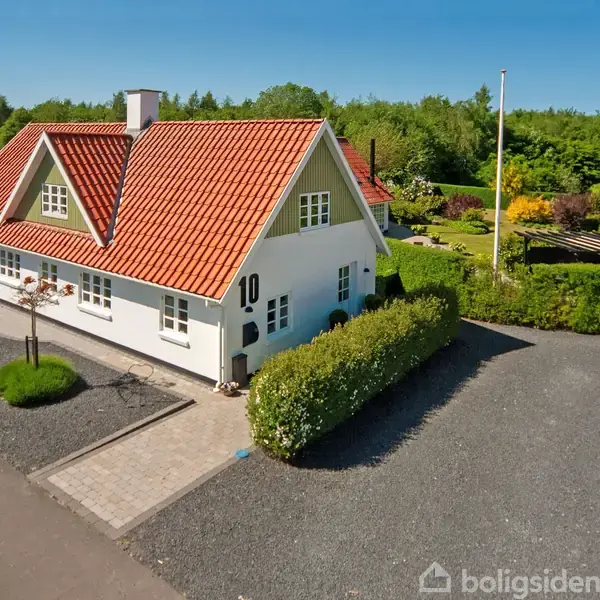 Huset er hvidt med rødt tegltag, står på en grusbelagt grund omkranset af grønne buske. Nummeret 10 er synligt på husets facade. I baggrunden ses træer og have.