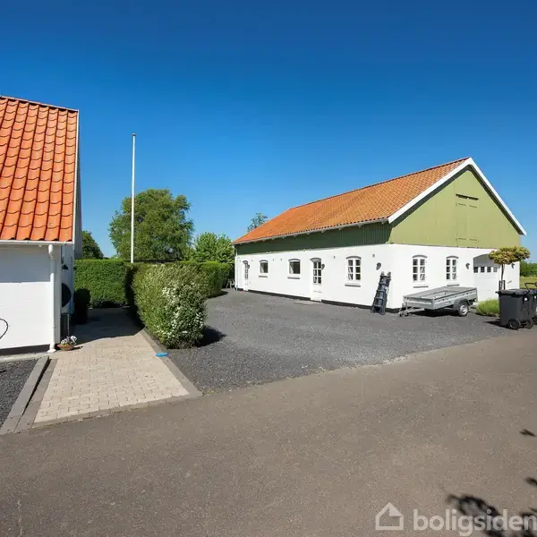 Hvid bygning med rødt tag står stille ved en asfalteret gårdsplads; omgivet af grønne træer og hække. Blå himmel i baggrunden, en trailer er placeret ved siden af bygningen.