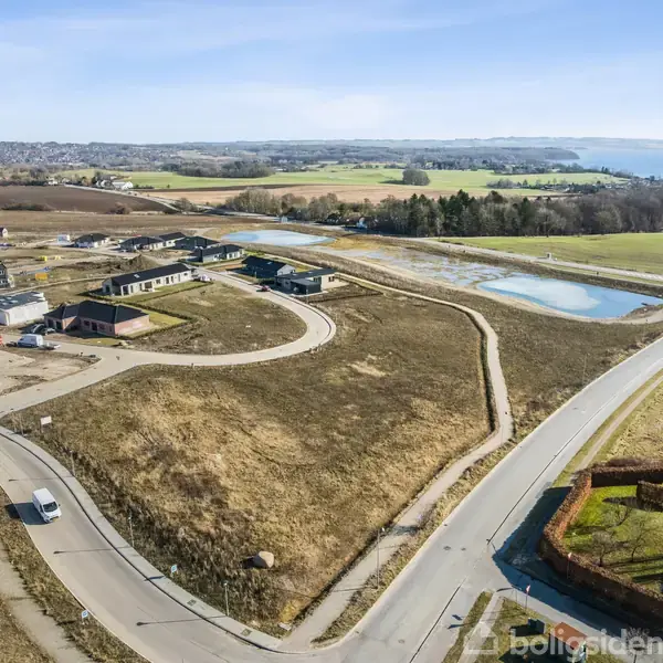 Boligområde under udvikling nær kysten, med spredte huse og byggeri. Krogede veje snor sig gennem området, omgivet af åbne marker og et lille vandreservoir. I baggrunden ses havet og skovområder.