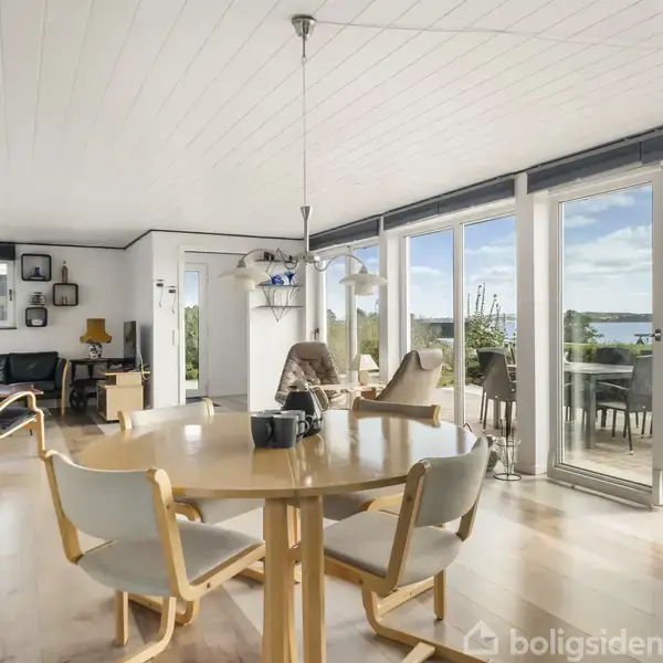 Rundt spisebord med træstole står i en lys stue. Store vinduer og skydedøre fører ud til en terrasse med udsigt over vandet. Møbler omfatter sofa og lamper.
