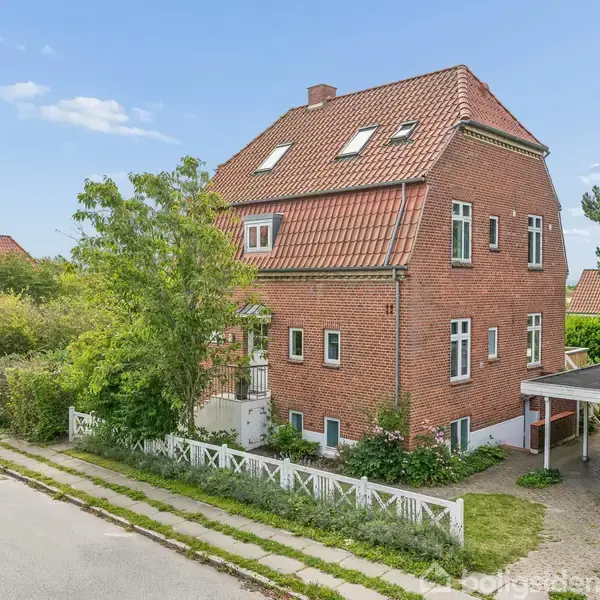 En murstensbygning med skråt rødt tegltag står stille omgivet af frodige træer og en hæk. Foran er der en carport ved en rolig vej.