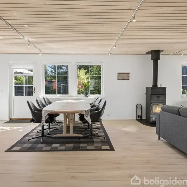 Stue med spisebord omgivet af stole, placeret på et tæppe. En brændeovn står ved siden af sofaen. Store vinduer og havedør slipper dagslys ind. Plankegulv og træloft.