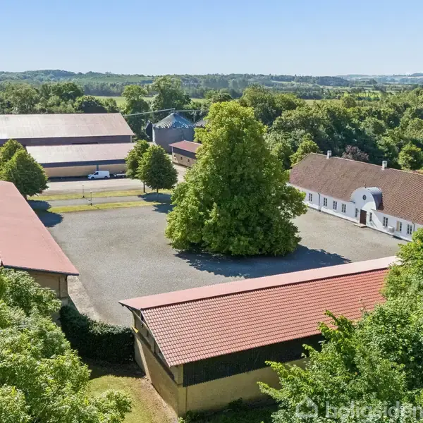 En stor gård ligger omkranset af flere rødtage bygninger, med en gruset gårdsplads i midten og frodig grøn bevoksning omkring. En stor træ står centralt på pladsen.