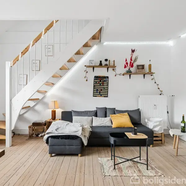 En grå sofa med gule puder og tæppe står ved en hvid væg med trin op ad en trappe. Et vinreol er monteret ved siden af sofaen i stuen.