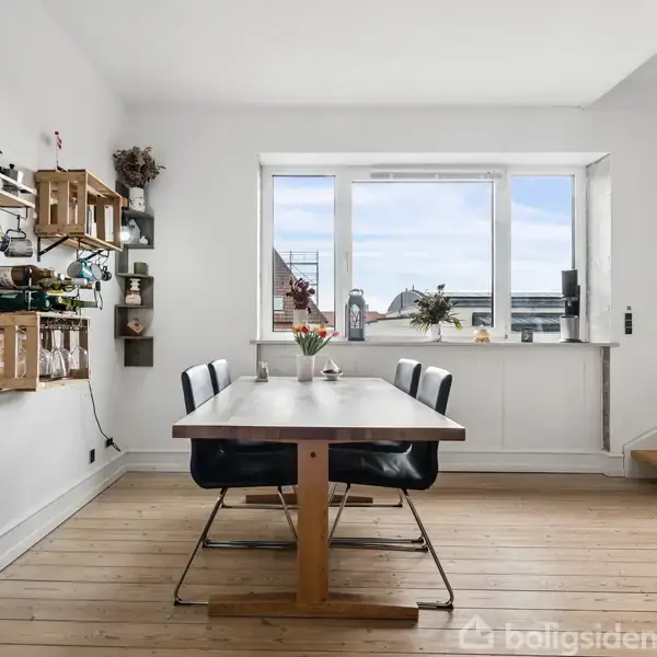 Spisebord står i en lys stue ved et stort vindue. Fire sorte stole omgiver bordet, og der er hylder med planter og dekorationer på væggen.